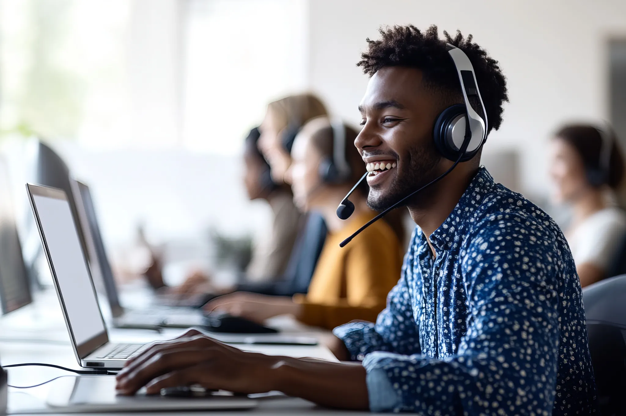 Smiling customer service representative wearing a headset, working on a laptop in a modern office with colleagues in the background.