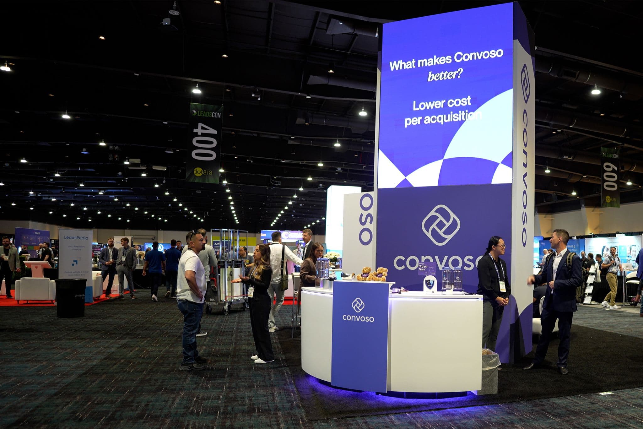 People interacting at a trade show booth for Convoso, featuring a display with the text "What makes Convoso better? Lower cost per acquisition."