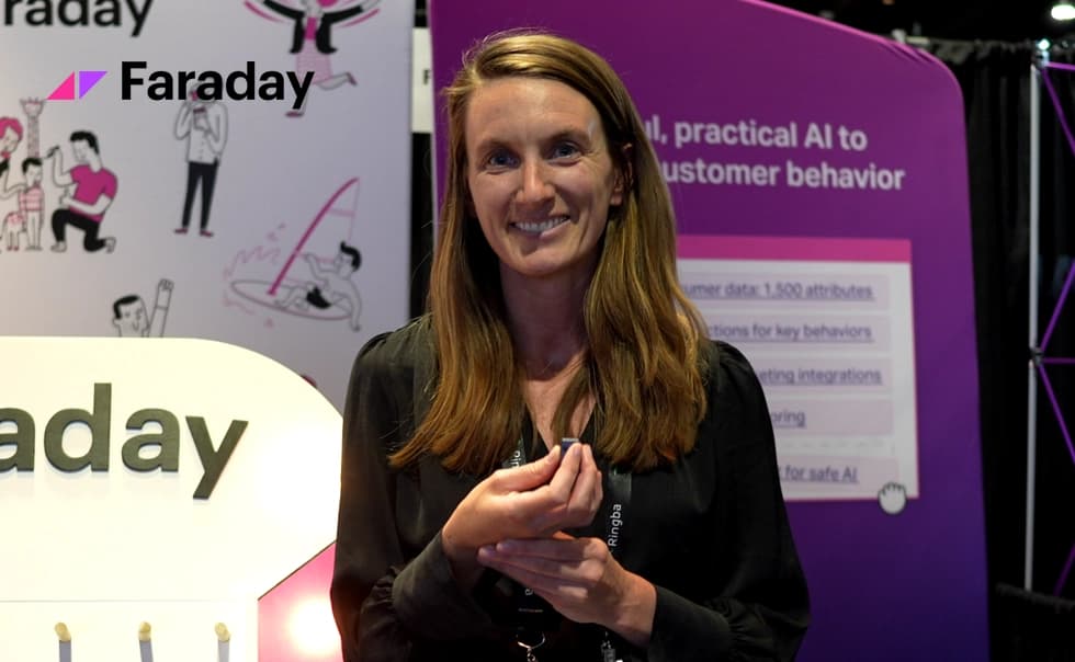 Smiling woman with long hair stands in front of a Faraday booth displaying AI solutions, featuring a purple backdrop with text and illustrations.