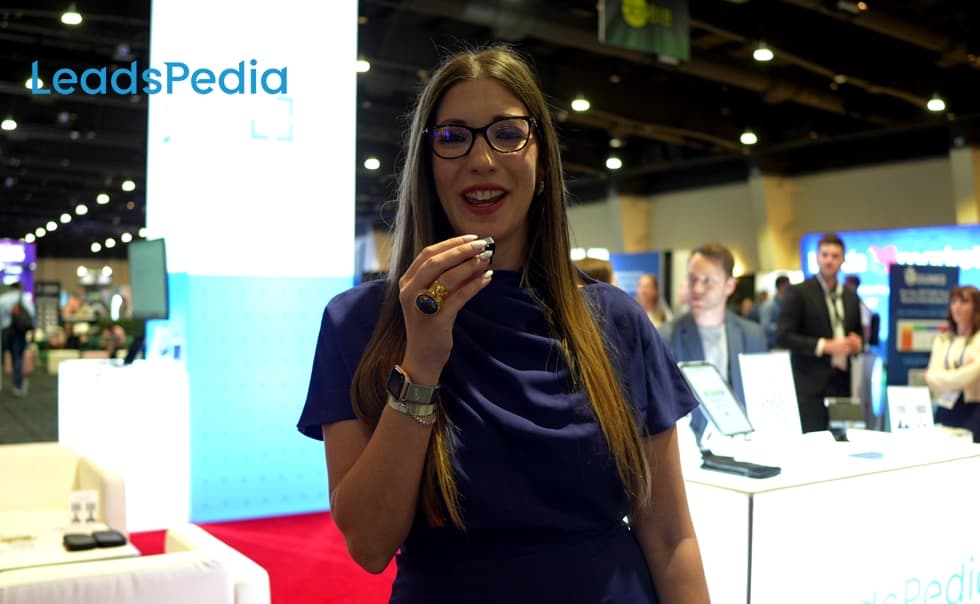 A woman in glasses and a blue dress speaks into a microphone at a tech conference. Booths and attendees are visible in the background.