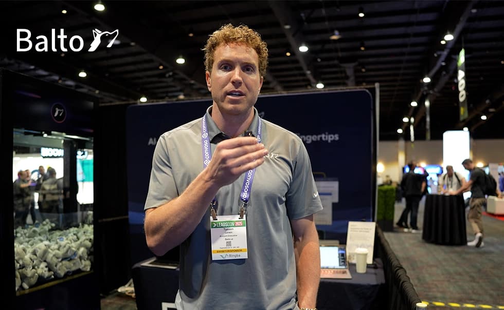 Man speaking at a convention booth with "Balto" logo, wearing a gray shirt and conference badge, standing in a well-lit exhibition hall.