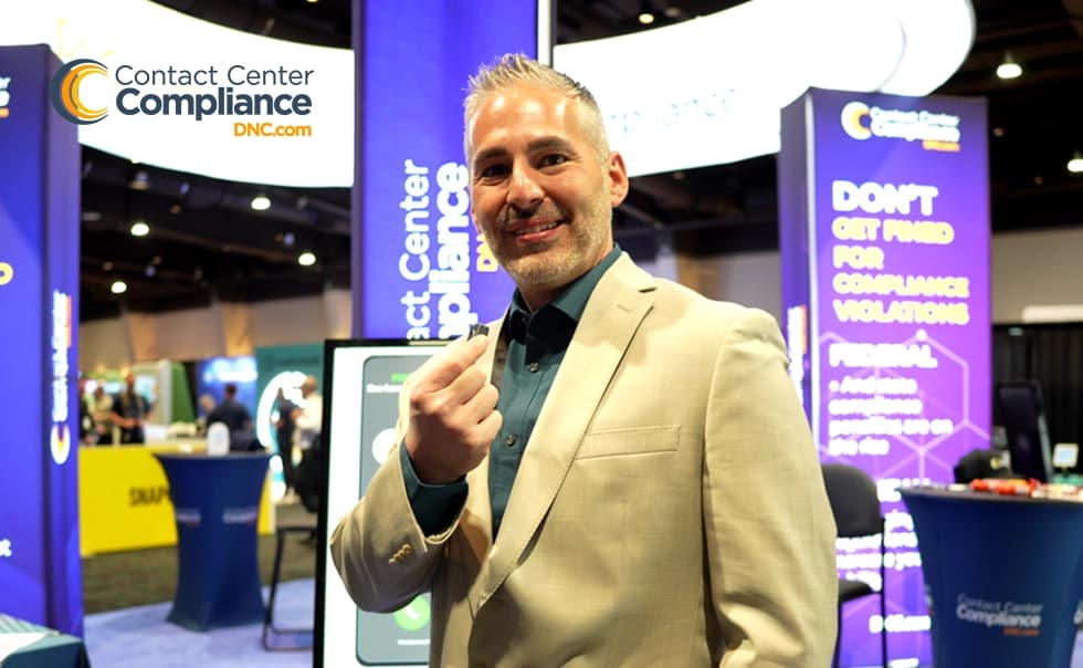 A man in a beige suit smiles and gestures in front of a trade show booth for Contact Center Compliance.