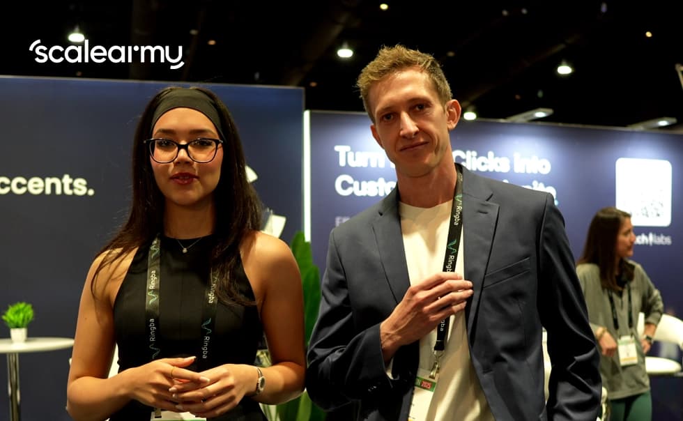 Two people stand at a conference booth with "scalearmy" branding. They wear lanyards and are in a professional setting.