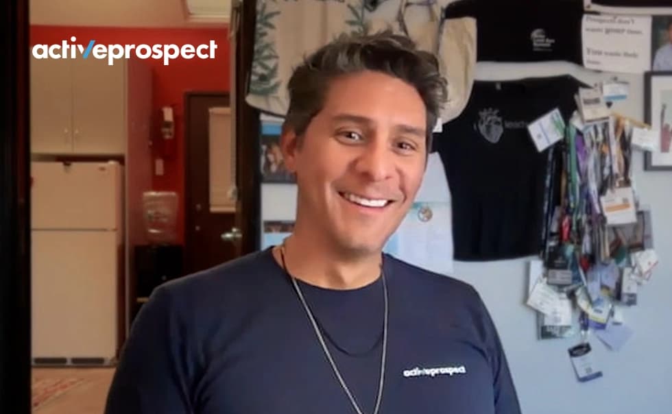 Smiling person wearing a navy "activeprospect" shirt, standing in a room with various items on the wall.