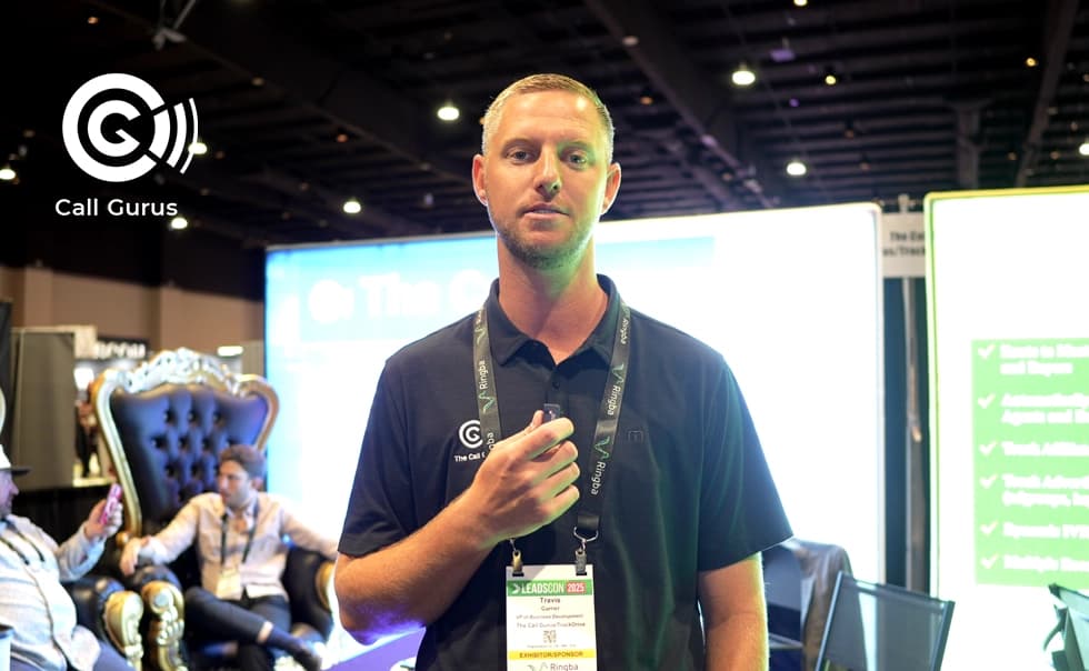 Man in a black Call Gurus shirt holding a microphone at a convention booth, with a large chair and two people in the background.