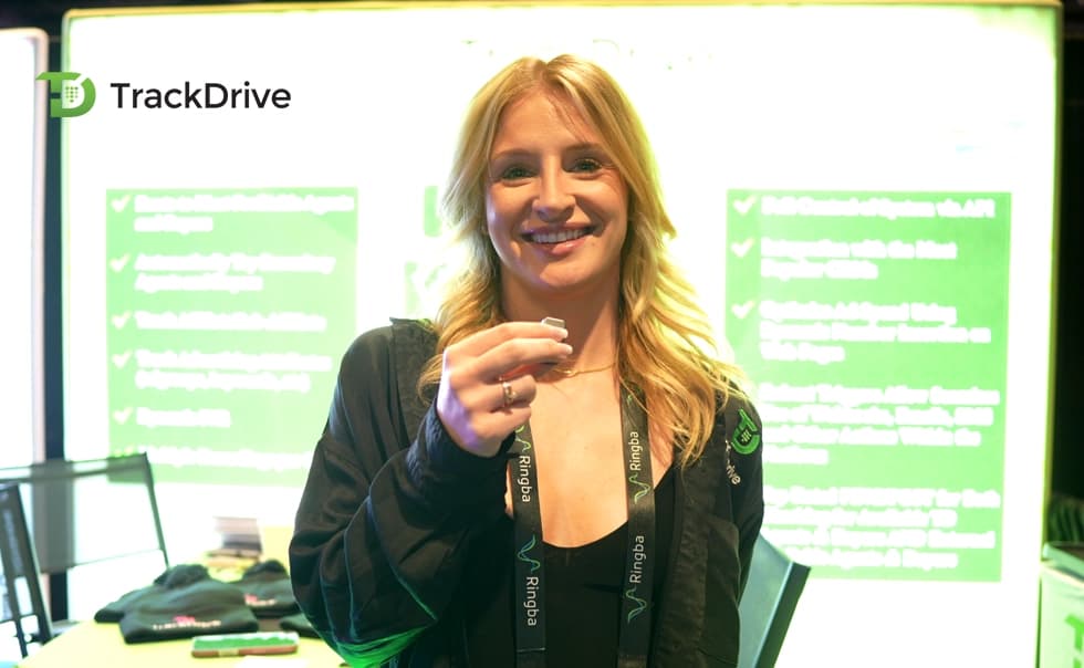 Smiling person holding a small object, standing in front of a bright green display with text. TrackDrive logo visible in the top left corner.