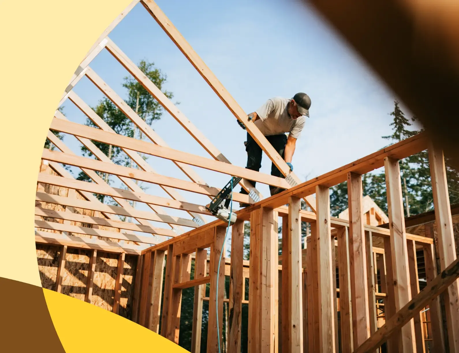A construction worker wearing a cap and gloves works on the wooden frame of a building under a clear blue sky.