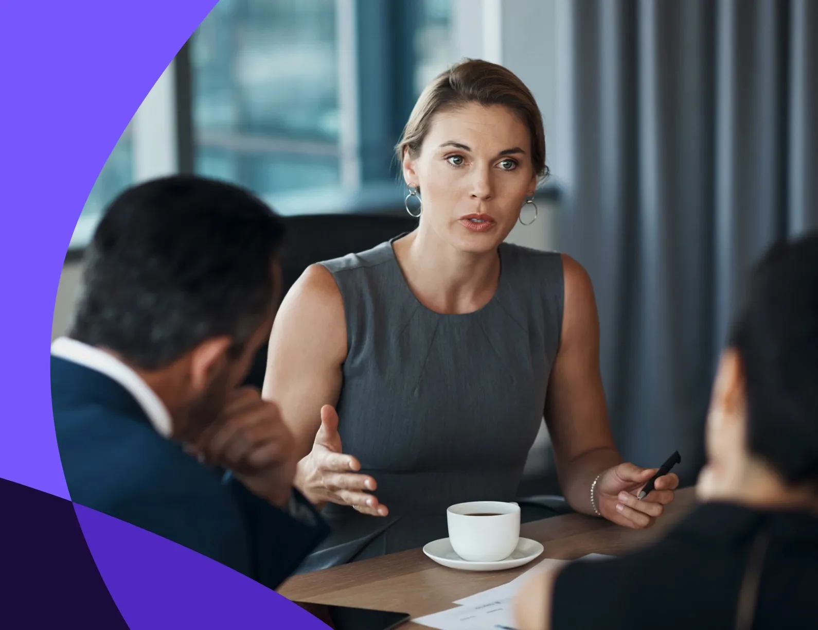 A woman in a gray dress speaks seriously at a meeting table with two colleagues, holding a pen, with a cup of coffee in front of her.