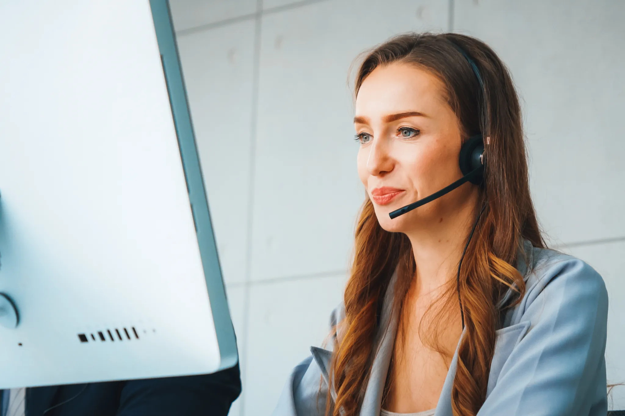 Woman with long hair wearing a headset, working at a computer in a modern office setting.