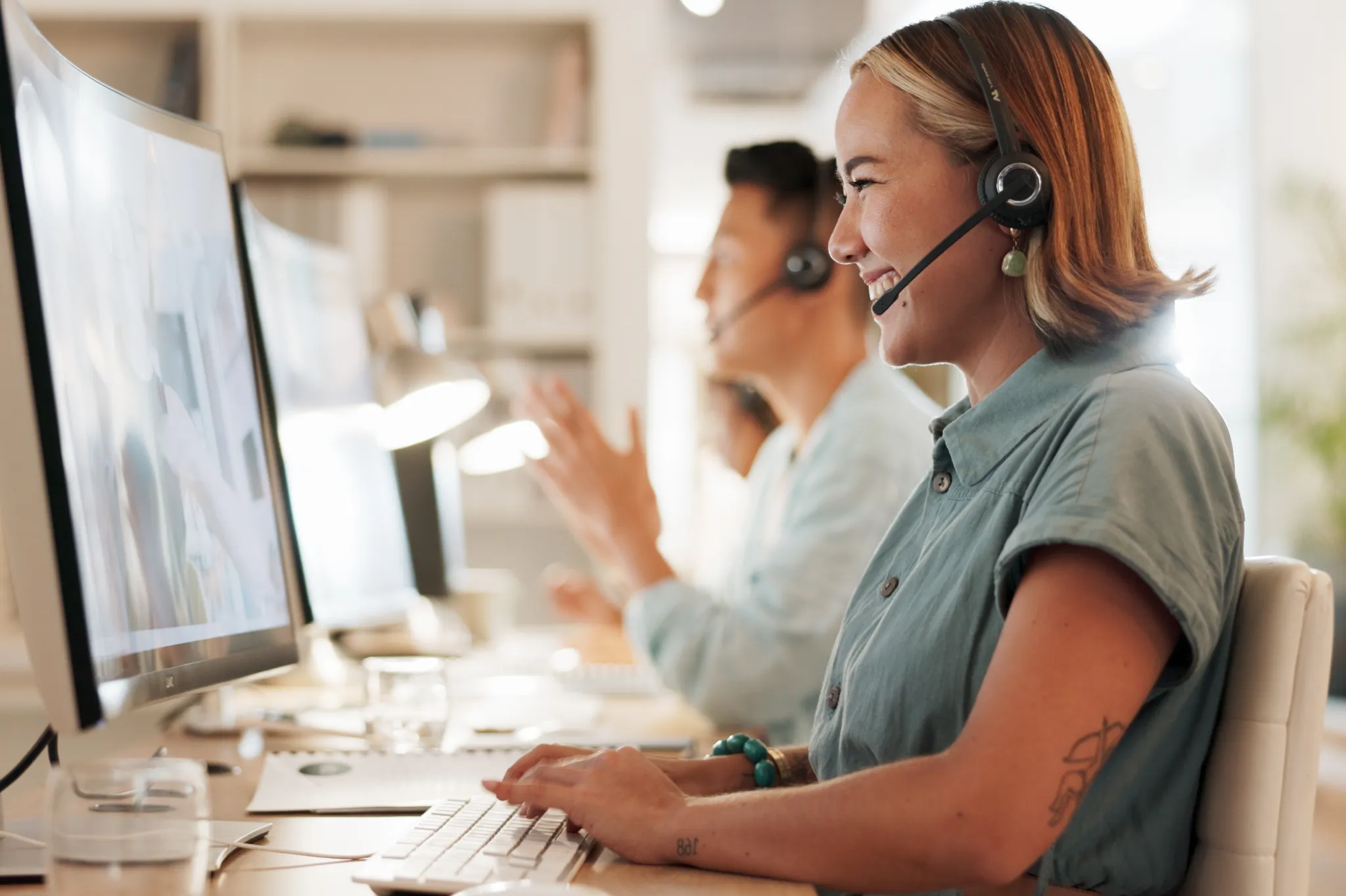 A woman wearing a headset smiles while working at a computer in an office. Other colleagues are visible in the background, also wearing headsets.