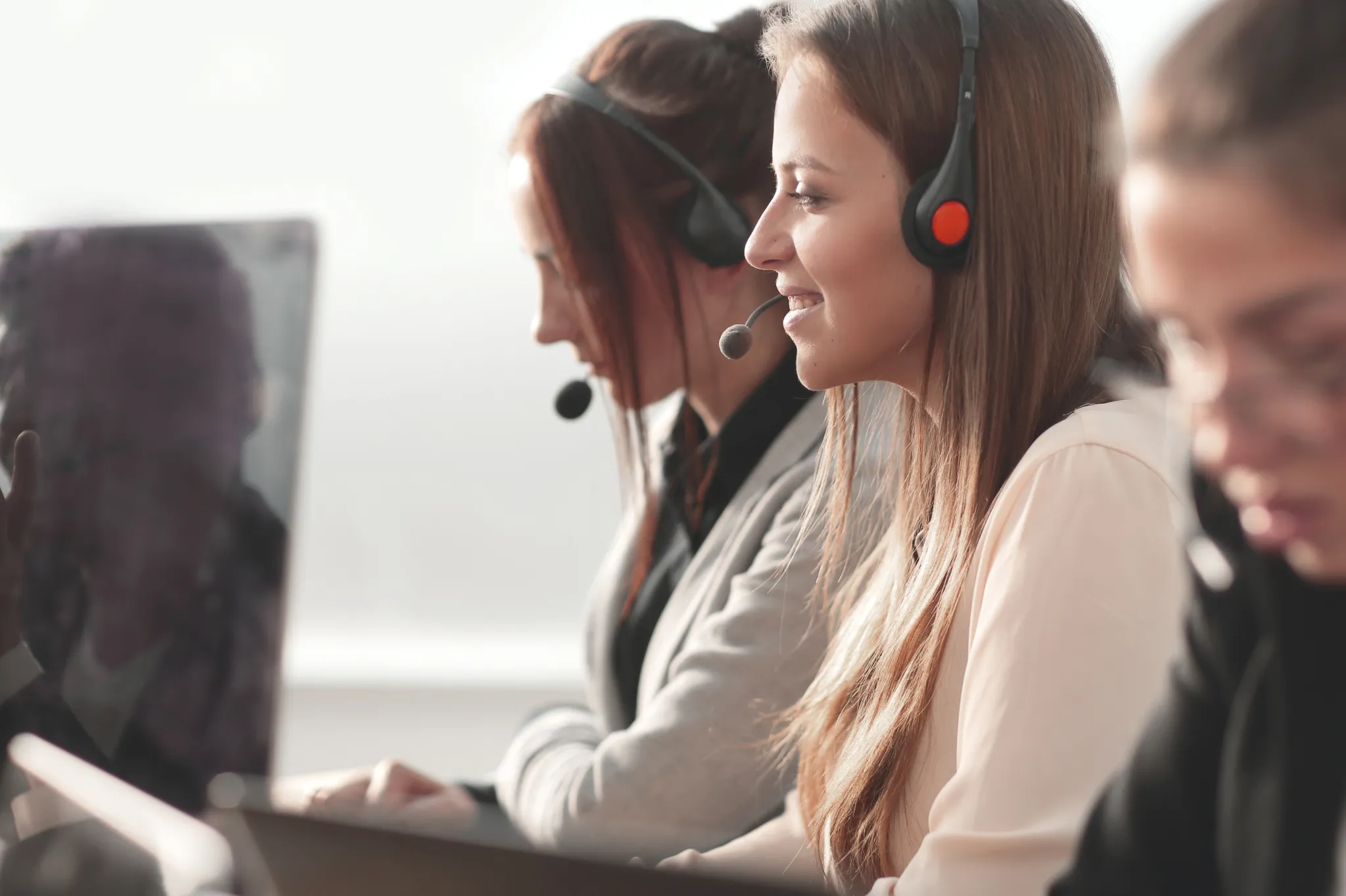 Three women wearing headsets work at computers in a bright office, focusing on tasks, with one smiling in the foreground.