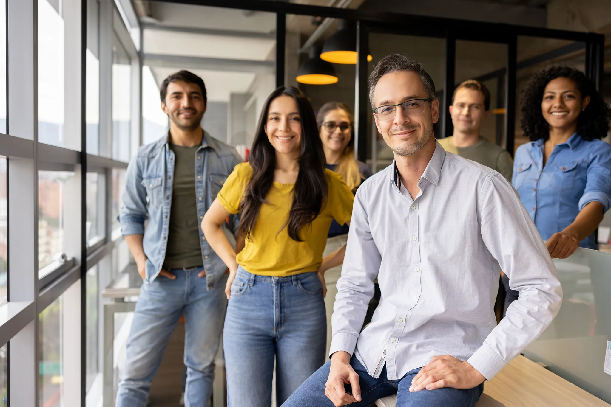 A group of six people smiling in an office near large windows, with one person sitting on a desk in front.