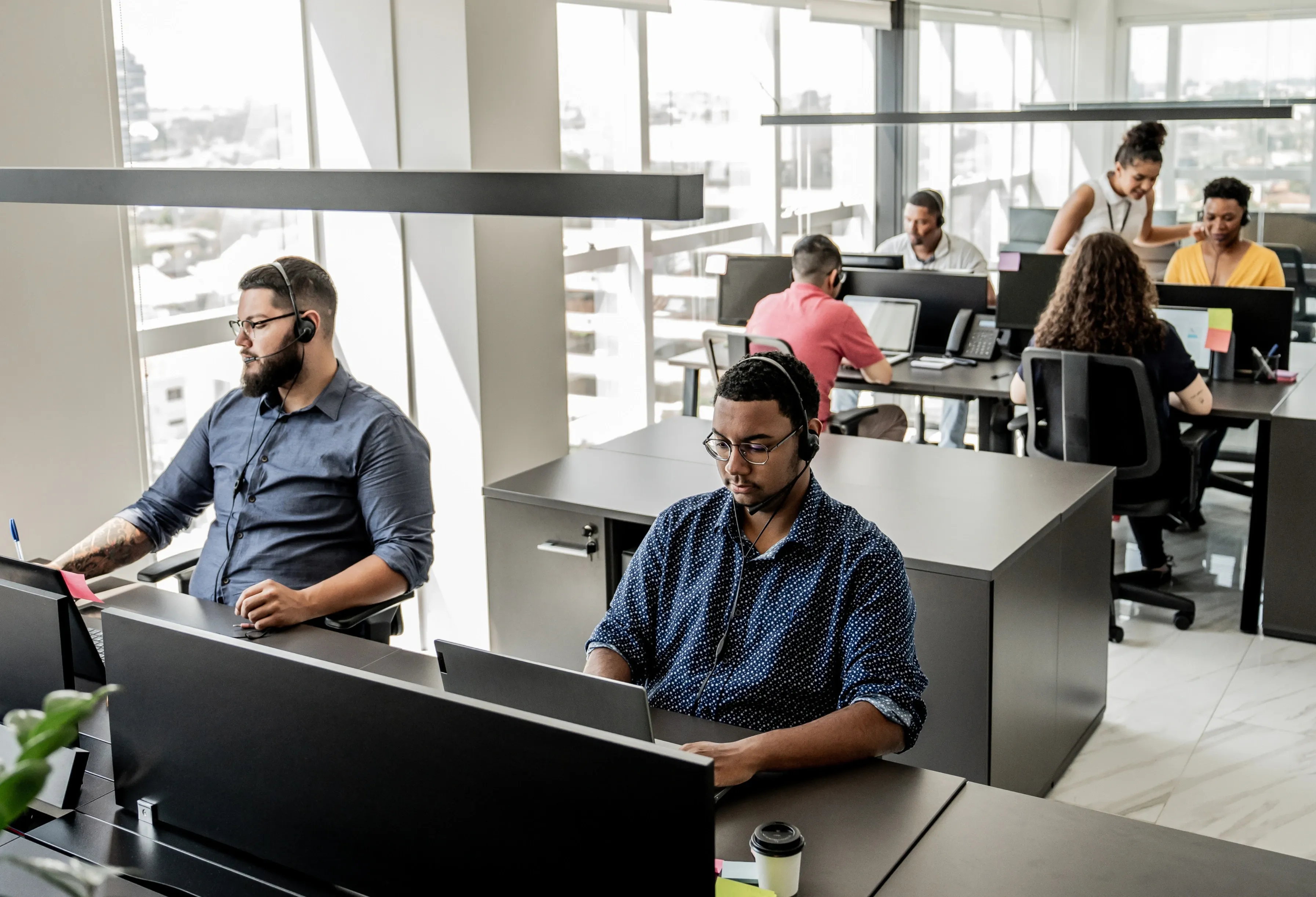 Open-plan office with diverse employees working at computers, wearing headsets. Bright space with large windows and modern furniture.